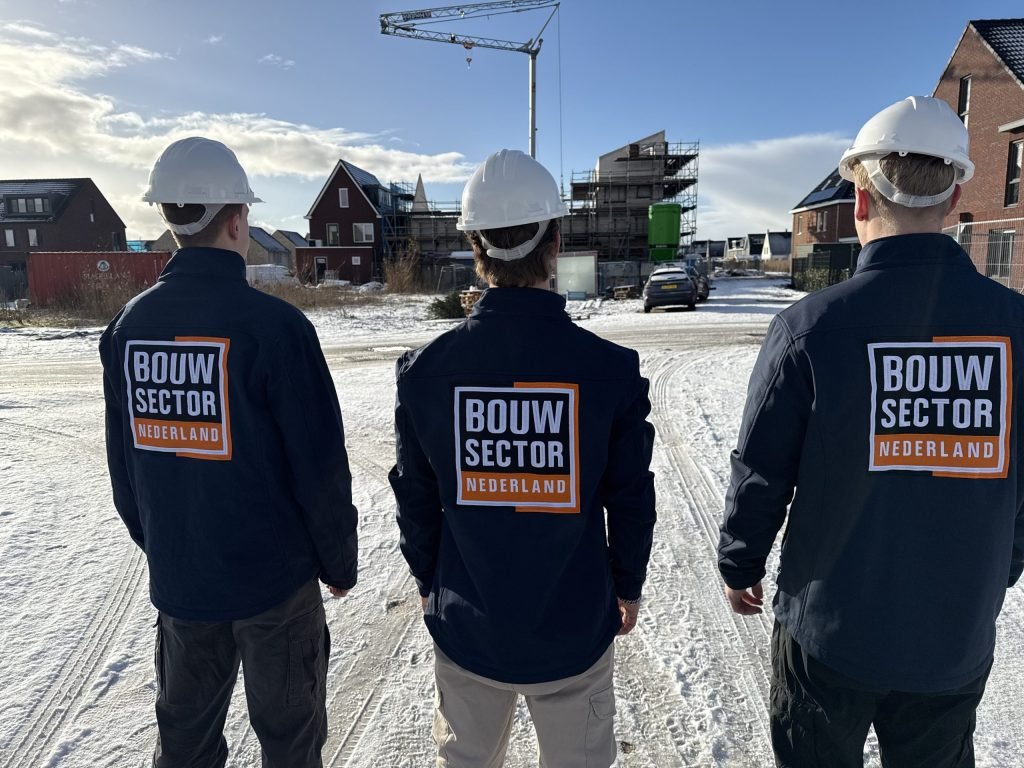 Drie vakmensen van Bouwsector Nederland met witte helm kijken naar huizen in aanbouw in een besneeuwde nieuwbouwwijk
